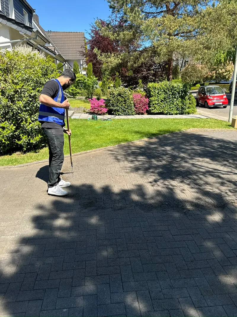 Mitarbeiter in Warnweste entfernt mit einem Fugenkratzer Unkraut aus dem Pflaster einer sonnigen Hofeinfahrt.