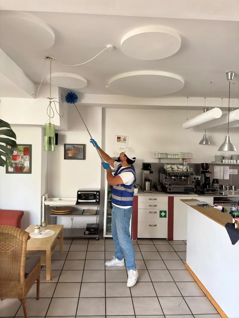 Mitarbeiter entstaubt mit einem Teleskopwedel die Deckensegel in einem Gastraum mit Theke und Kaffeemaschine.