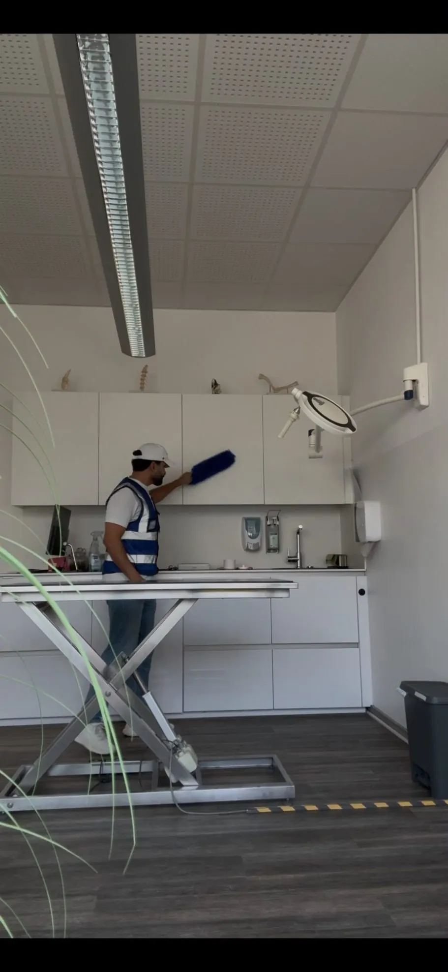 Mitarbeiter entstaubt mit einem Wischer die Oberkanten der Hängeschränke in einem Behandlungsraum.